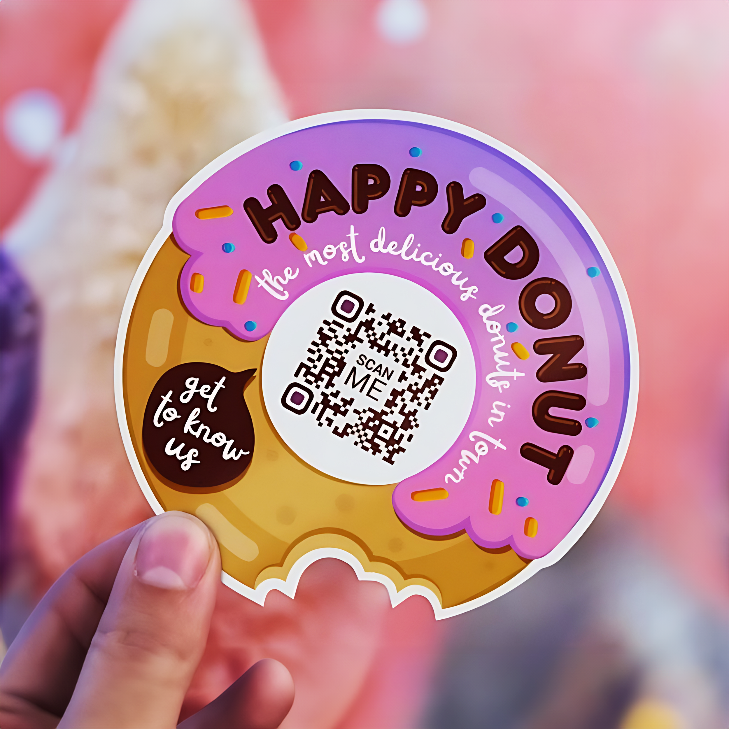 The image shows a person holding a sticker that resembles a donut with a bite taken out of it. The sticker features the text "HAPPY DONUT" in large, bold letters at the top, with a tagline below that reads "the most delicious donut in town." There is a QR code in the center with the text "SCAN ME" beneath it. To the left, there is a small text bubble that says, "get to know us."